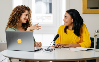 Two women sit around a circular table to record a podcast. The woman on the left is sitting in front of a laptop, and the woman on the right is behind a microphone.